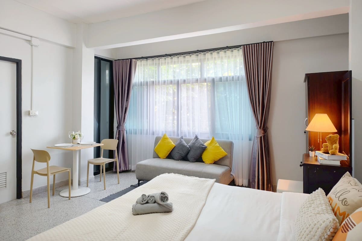 Habitación de hotel con cama, escritorio, silla, sofá con cojines, ventana con cortinas y lámpara.