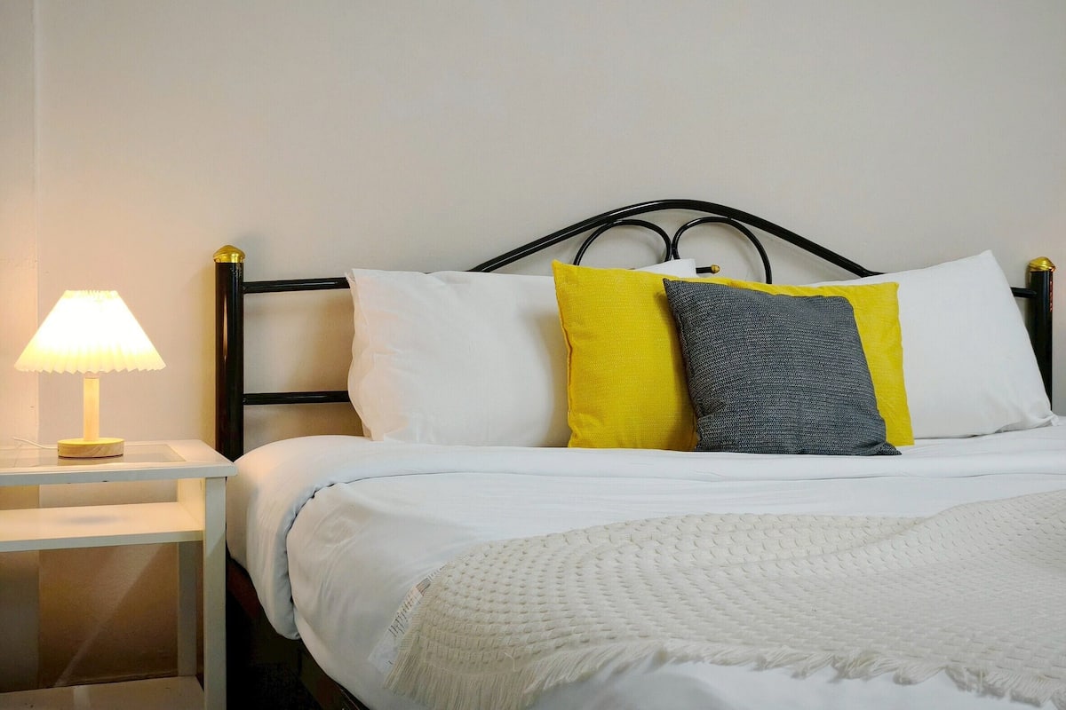 Una cama con ropa de cama blanca, un cabecero de metal negro y una mesita de noche con una lámpara.