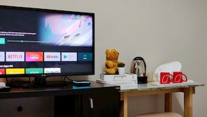 A television displaying a movie scene, a teddy bear, a coffee pot, and a desk with a chair and red mugs. - Xin Hotel at Udomsuk 29 (Bangkok)