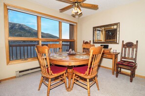 A dining room with a round wooden table, six chairs, a ceiling fan, large windows with a view of mountains, and a mirror on the wall.