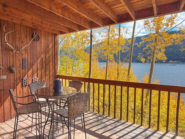Um deck de madeira com mesa e cadeiras, com vista para um lago cercado por folhagem amarela e montanhas.