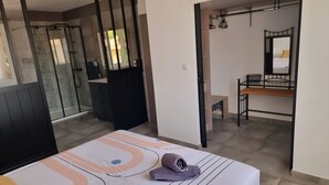 A bedroom with a bed, a bathroom with a shower, and a small sitting area with a bench and table. - House in the heart of the Alpilles (Maussane-les-Alpilles)