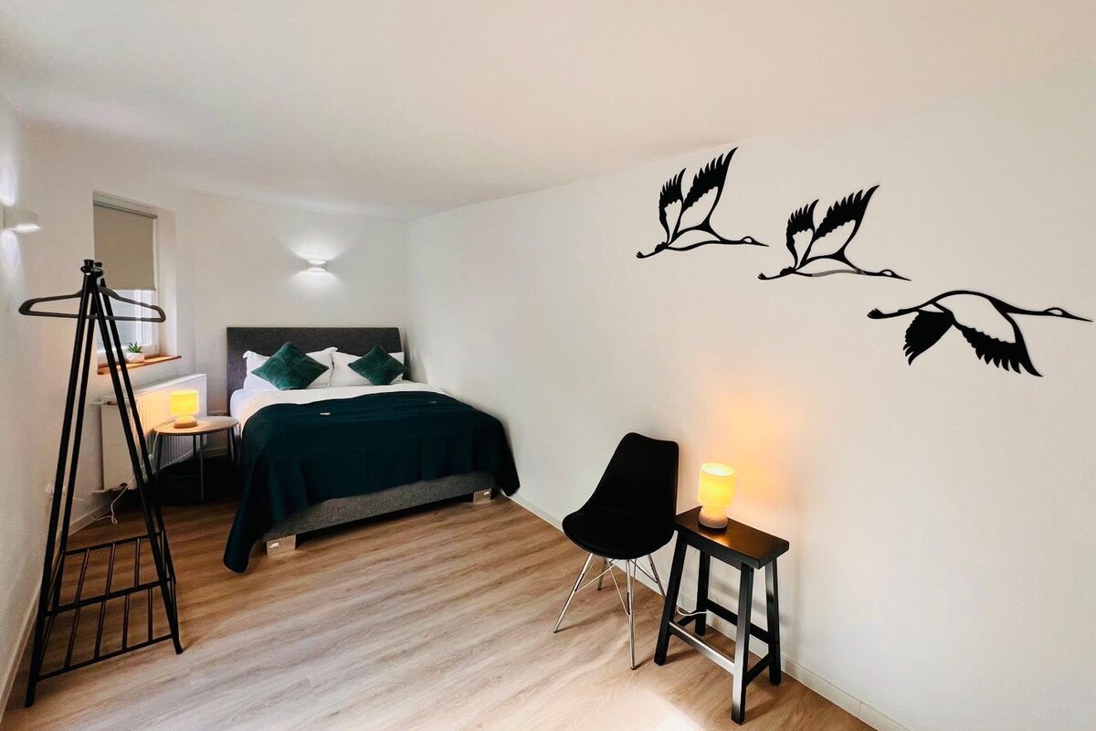 Un dormitori amb llit, taula de nit, cadira, làmpada i obra d'art a la paret.