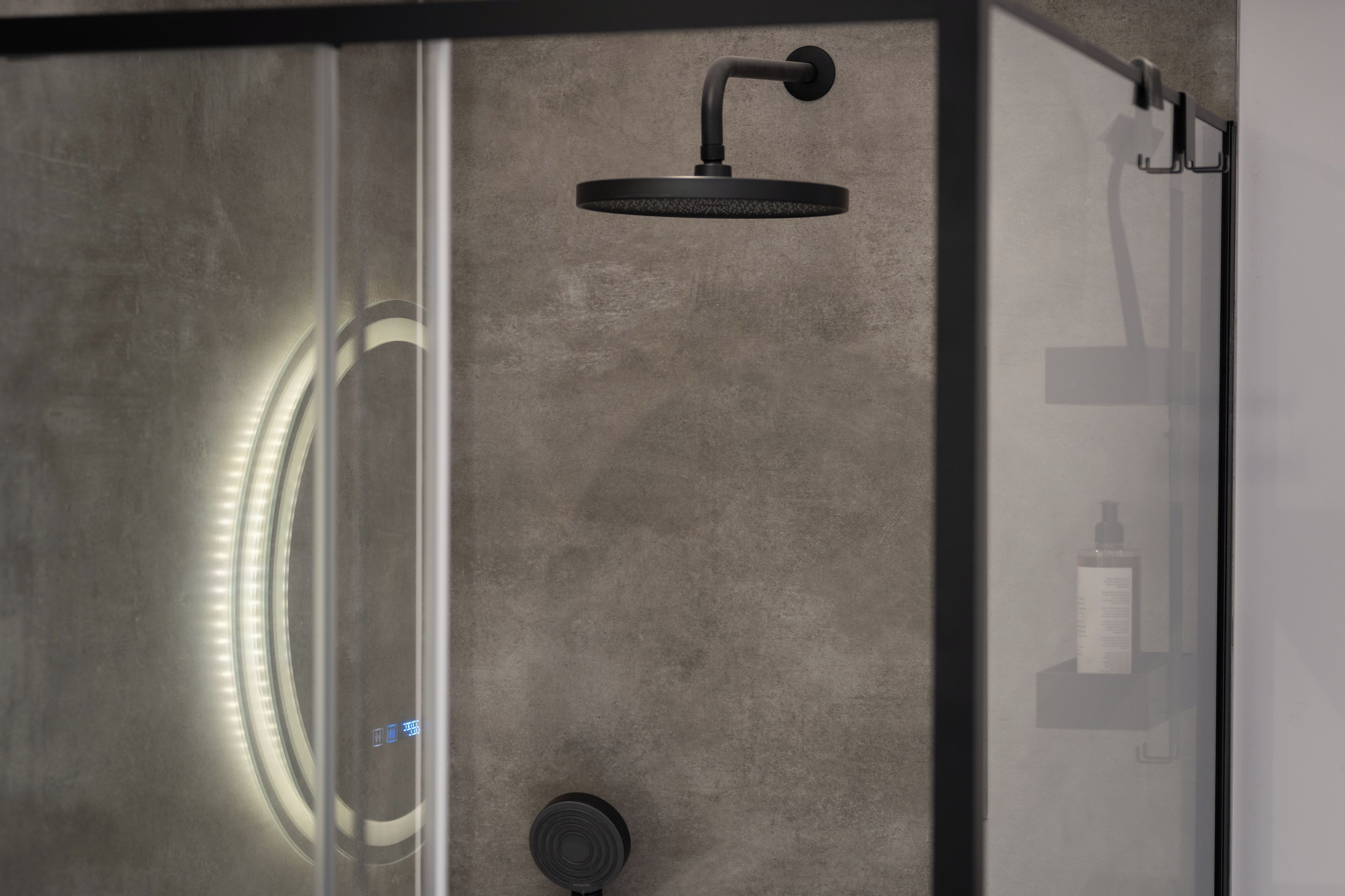 Une douche moderne dotée d’une pomme de douche incurvée, d’un espace clos en verre et d’une bouteille visible sur une étagère.