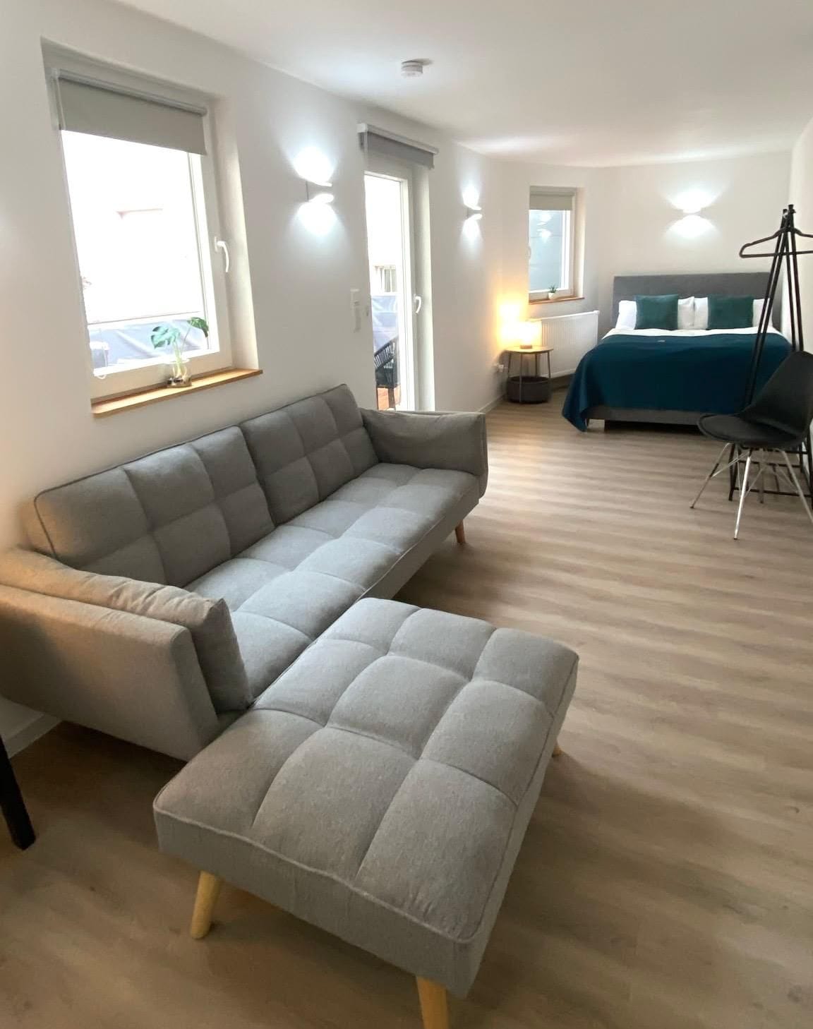 A modern living room with a grey sectional sofa, a small ottoman, a bed with a blue bedspread, and a chair.