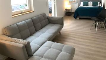 A modern living room with a grey sectional sofa, a small ottoman, a bed with a blue bedspread, and a chair.