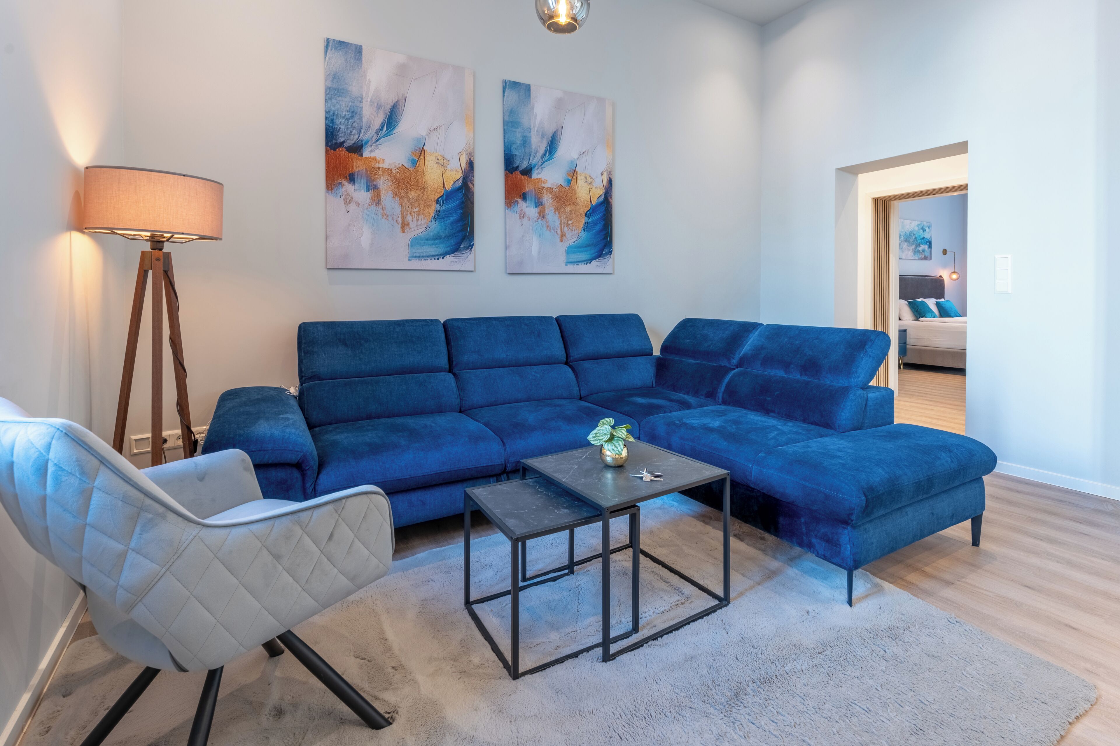 A living room with a blue sectional sofa, a small black coffee table, a white armchair, and two abstract paintings on the wall.
