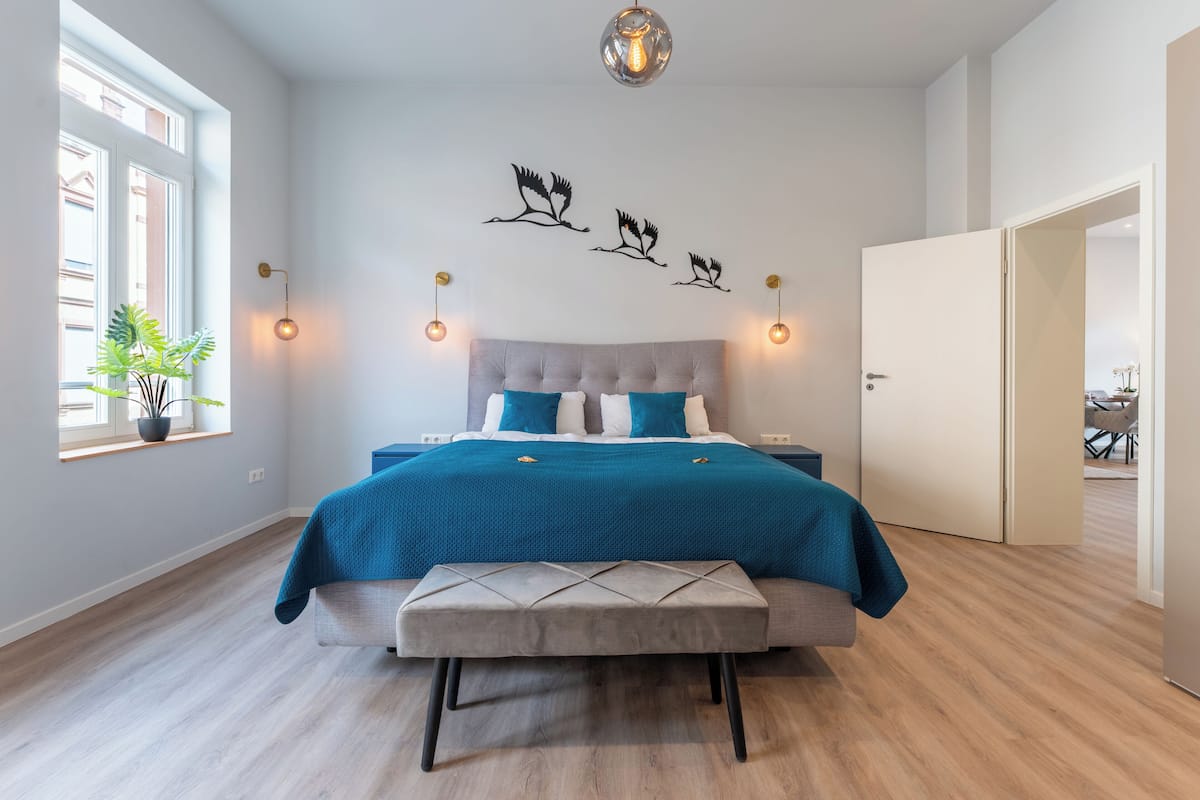 A bedroom with a bed, bench, wall art, and a window with a plant.
