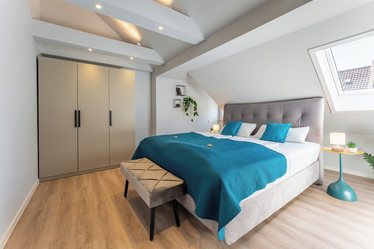 A bedroom with a bed, bench, wardrobe, bedside table, lamp, and a window.