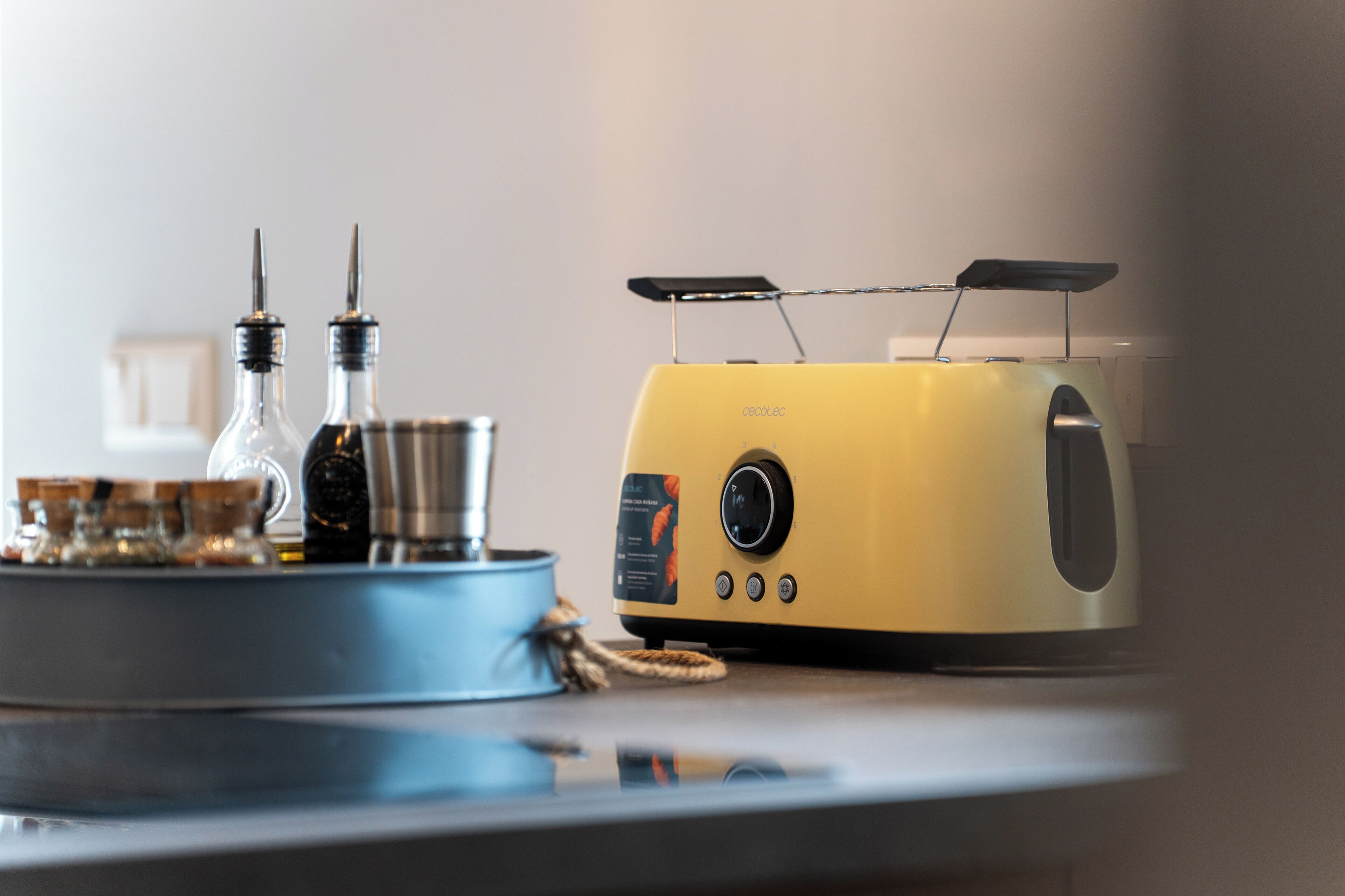Un grille-pain jaune doté d’un panneau de commande et de deux fentes, placé sur une surface réfléchissante avec un plateau bleu contenant un récipient en verre et un objet métallique.