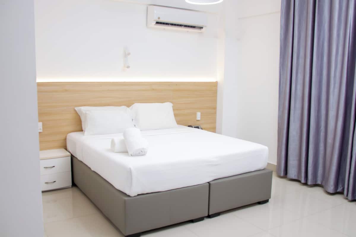 A neatly made bed with white linens, a grey headboard, and a small bedside table with drawers.