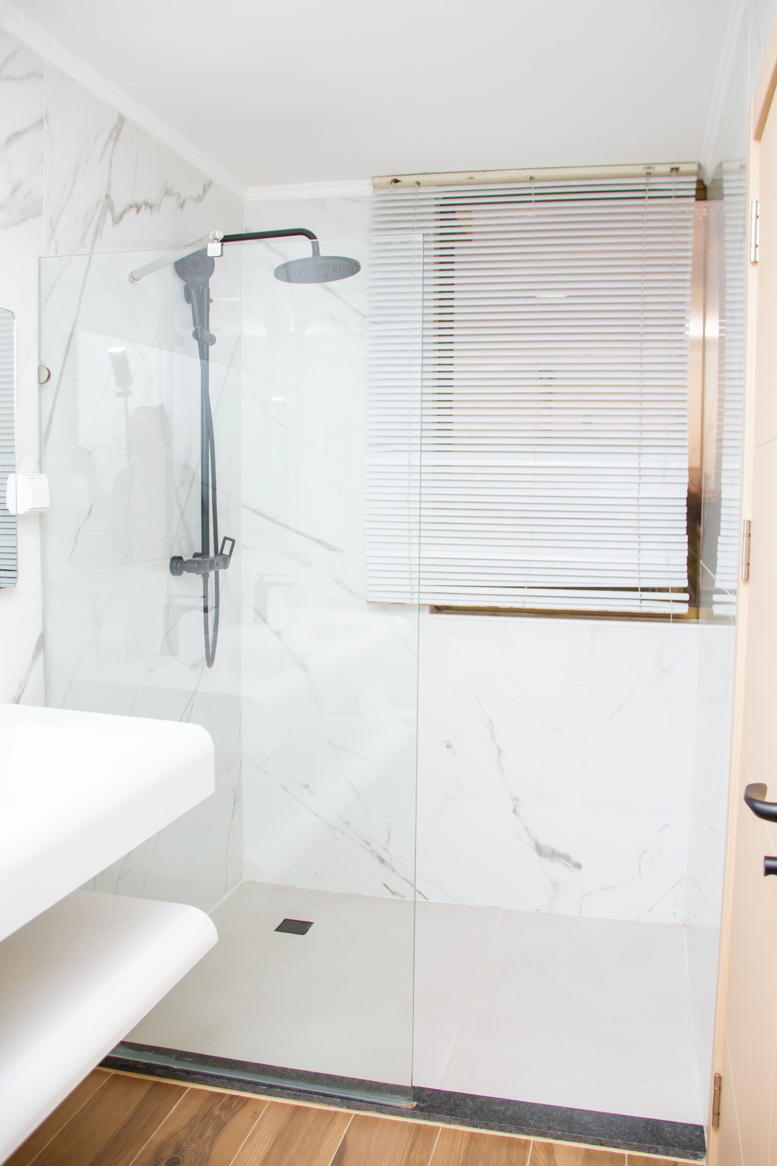 A modern bathroom with a glass shower enclosure, a white marble wall, and a wooden floor.