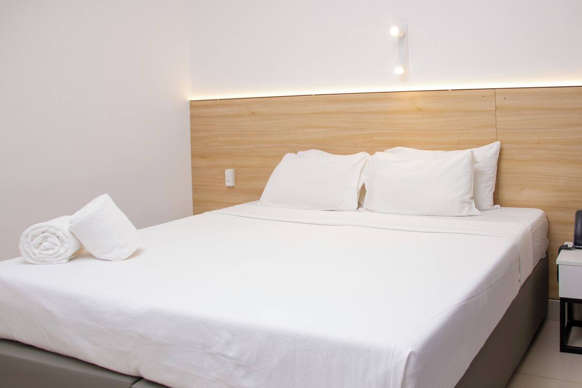 A hotel room with a neatly made bed, a rolled towel, a bedside table, and a wall-mounted light fixture.