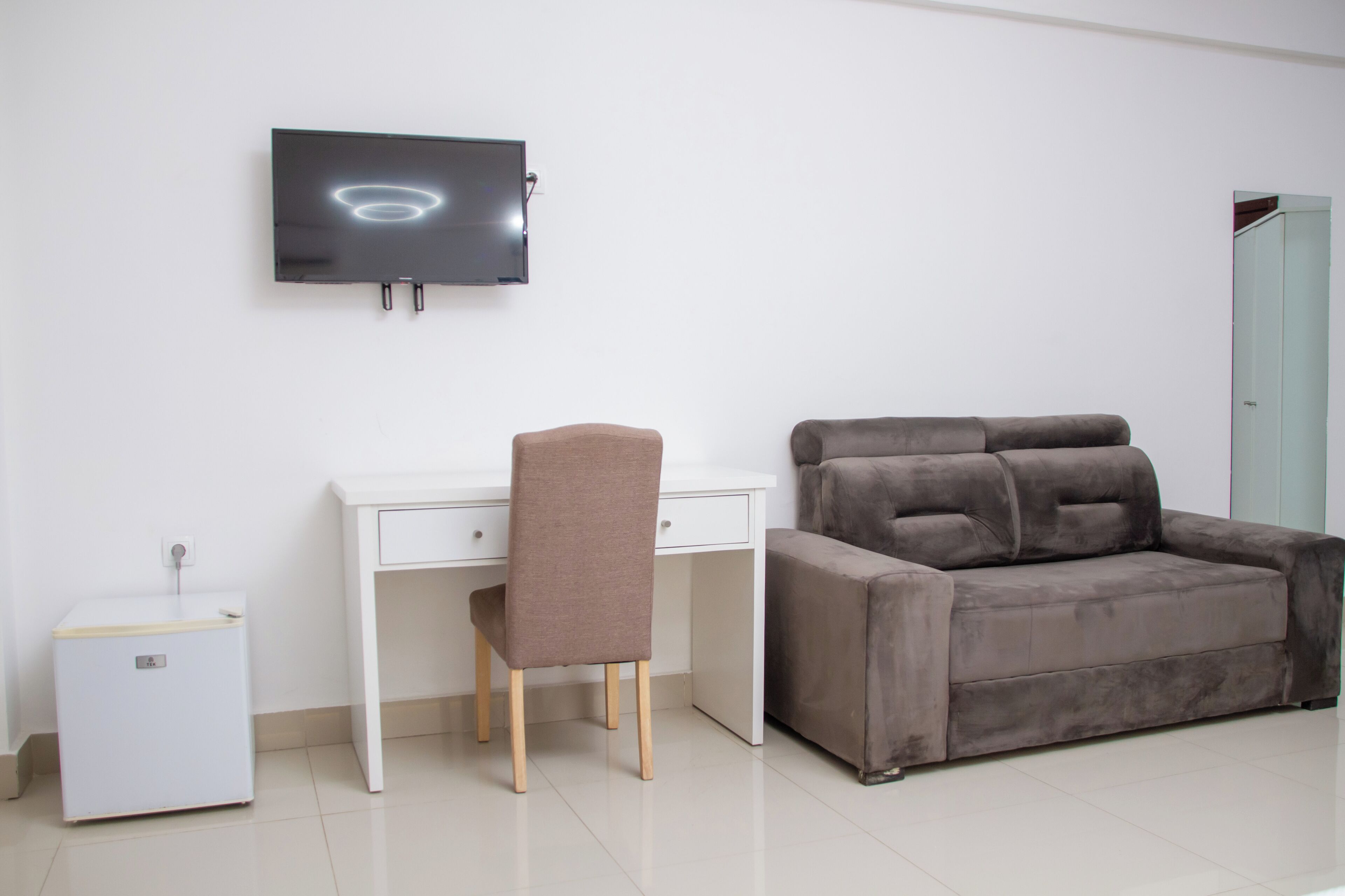 Une pièce comprenant un bureau blanc, une chaise marron, un canapé gris, un petit meuble blanc et un téléviseur à écran plat fixé au mur.
