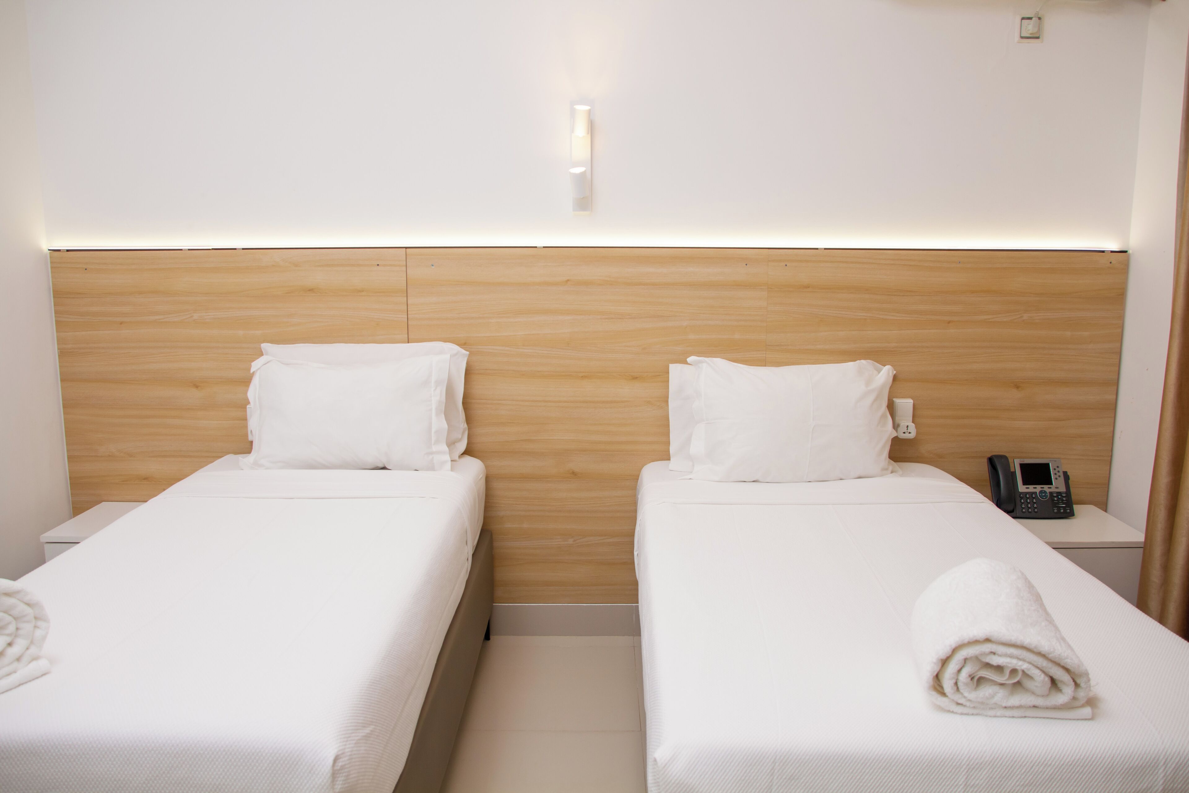 Habitación de hotel con dos camas individuales, cabecera de madera, teléfono en la mesita de noche y lámpara de pared.