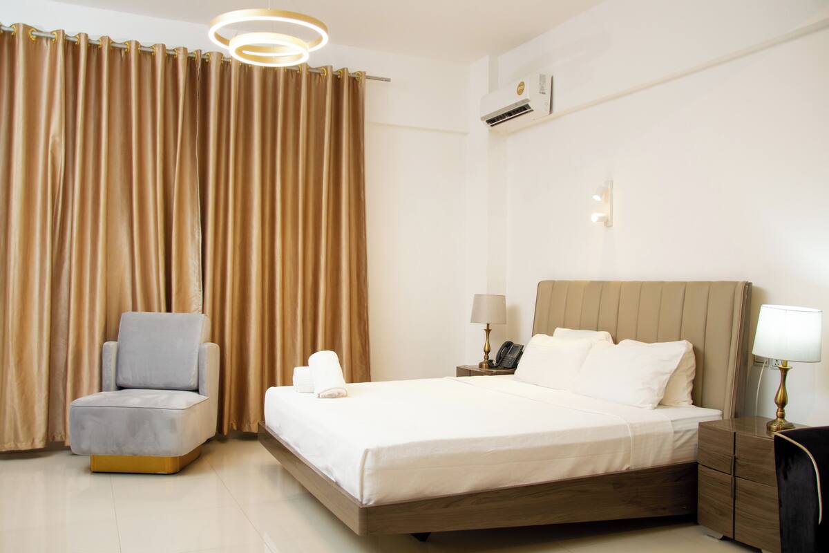 A hotel room with a bed, a chair, a bedside table with a lamp, and a curtain.