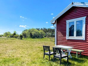 A red wooden cabin with a table and two chairs outside, surrounded by a grassy field and trees. - 4 Person Holiday Home in Borgholm (Borgholm)