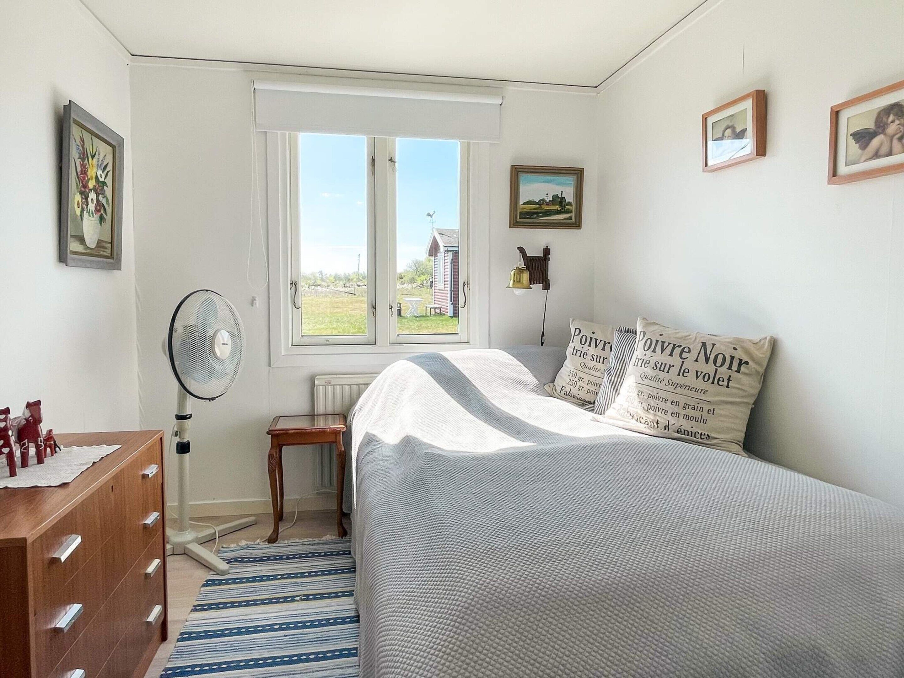 Quarto com uma cama, janela, radiador, cómoda de madeira e ventoinha.