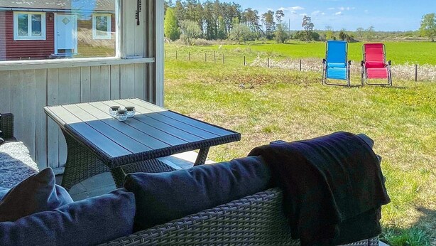 Ein überdachter Außenbereich mit Tisch und Korbmöbeln, mit Blick auf ein eingezäuntes Feld mit zwei bunten Stühlen.