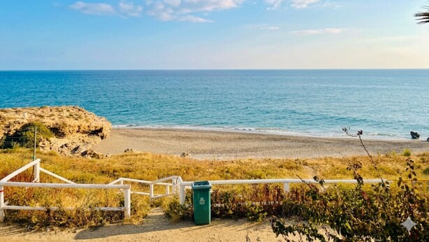 A beach with a white fence, a trash bin, and a clear view of the ocean. - Apartamento Sea Blue D D by At Home Costa del Sol (Torrox)