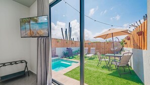 Ein modernes Wohnzimmer mit großem Fenster, einem Gemälde, einer Bank und Blick auf einen Garten mit Pool, Terrassenmöbeln und einem Holzzaun.