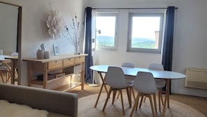 En moderne stue med sofa, spisebord med stole, et træskab og to vinduer med gardiner.