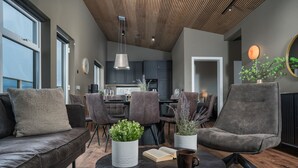 Un salon moderne comprenant un canapé, des fauteuils, une table basse avec des livres et des plantes, et donnant sur une cuisine et un coin repas.