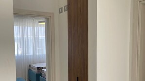 Un pasillo que conduce a un dormitorio con un gran armario de madera, una cama con cabecera blanca y una ventana con cortinas traslúcidas.