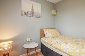 A bedroom with a bed, a lamp, a small table with a vase, and a cityscape painting on the wall. - Likely Sunset (St Austell)