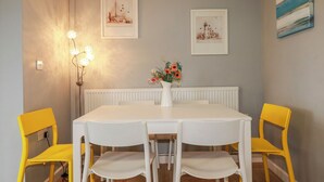 Un comedor con una mesa blanca, cuatro sillas, un jarrón con flores y dos cuadros en la pared.