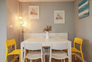 Un comedor con una mesa blanca, cuatro sillas, un jarrón con flores y dos cuadros en la pared.