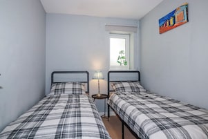 Un dormitorio con dos camas, una mesita de noche con lámpara, una ventana con vistas a zonas verdes y un cuadro en la pared.