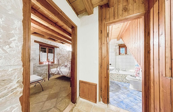 Ein rustikales Zimmer mit Steinwand, Holzbalken, einem Bett und Blick auf ein Badezimmer mit Badewanne und Toilette.