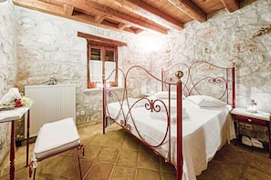 Une chambre avec un mur en pierre, un plafond en bois, un cadre de lit en métal, une chaise blanche et une petite table de chevet.