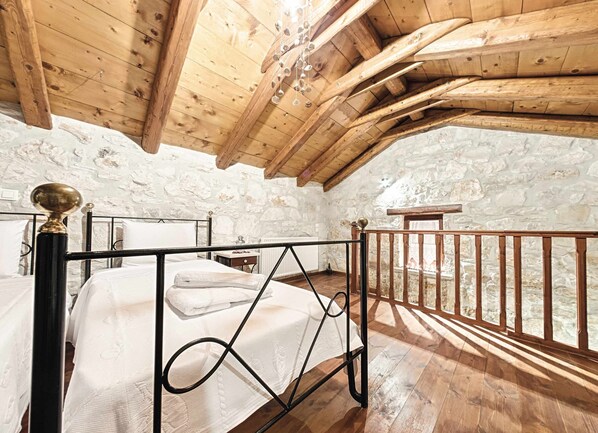 Une chambre avec un plafond en bois, des murs en pierre, un lit avec un cadre en métal et une balustrade en bois donnant sur une façade en pierre.