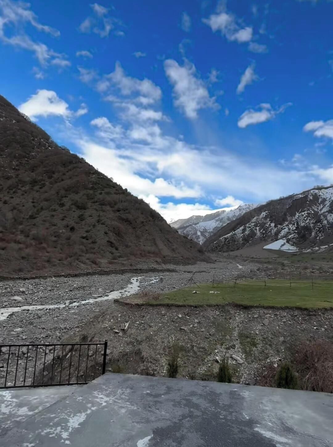 Un paisaje montañoso con un río, cielo despejado y una zona vallada en primer plano.