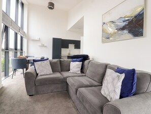 A modern living room with a grey sectional sofa, a dining table with chairs, a kitchen in the background, and a large abstract painting on the wall. - Apartment 20, River View (Preston)