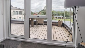 A room with large windows, a wooden deck, and a view of buildings and greenery. - Apartment 20, River View (Preston)