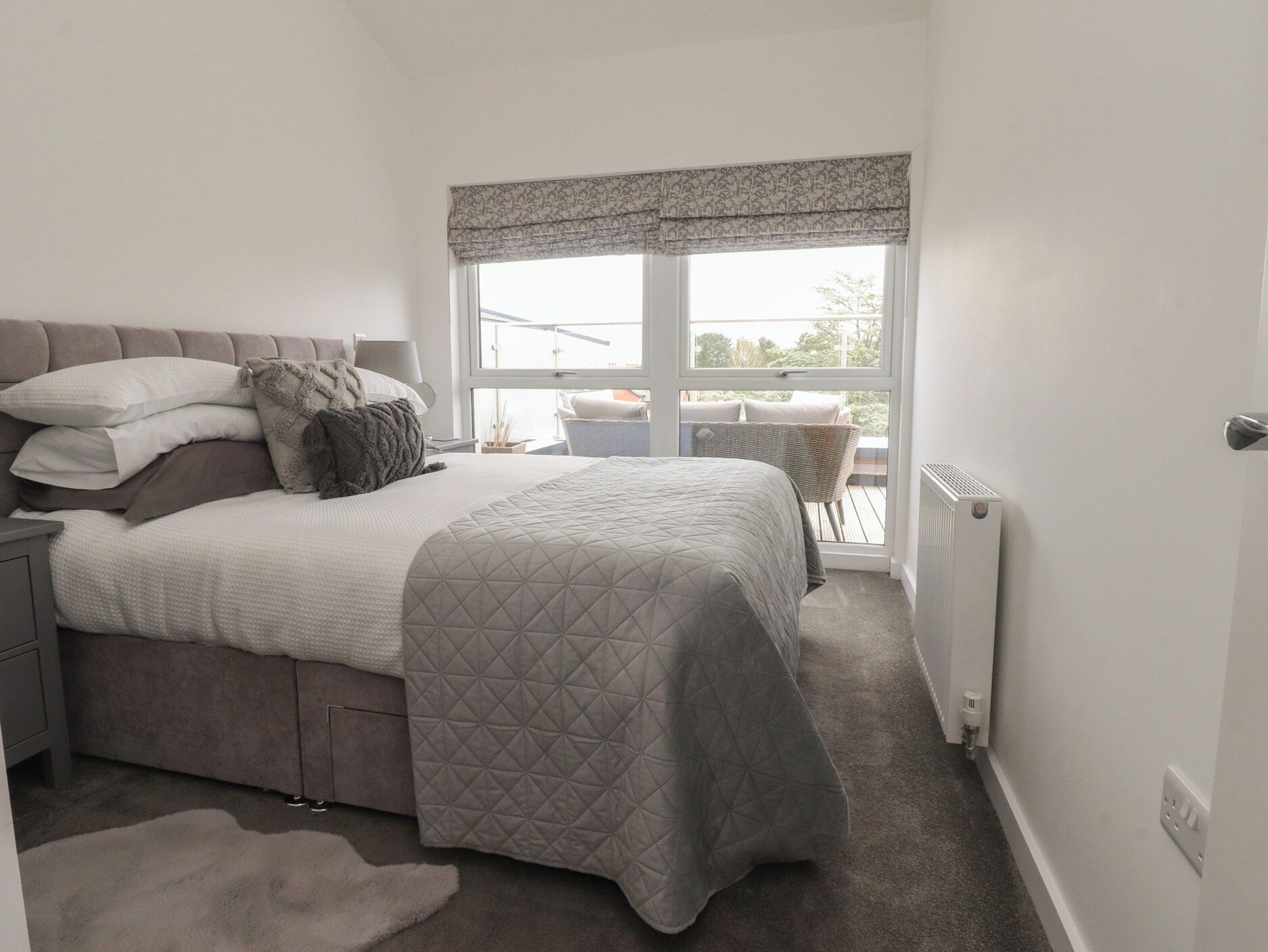 Une chambre à coucher avec un lit, des tables de chevet, un radiateur, une fenêtre donnant sur un beau paysage et un fauteuil en osier.