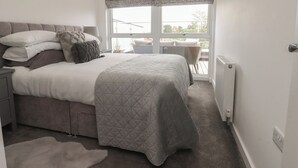 A bedroom with a bed, bedside tables, a radiator, a window with a view, and a wicker chair. - Apartment 20, River View (Preston)