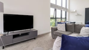 A modern living room with a flat-screen TV on a grey entertainment unit, a grey sofa with blue and white cushions, and a large window providing a view of greenery outside. - Apartment 20, River View (Preston)