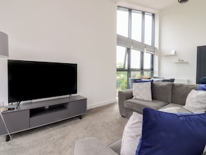 A modern living room with a flat-screen TV on a grey entertainment unit, a grey sofa with blue and white cushions, and a large window providing a view of greenery outside. - Apartment 20, River View (Preston)