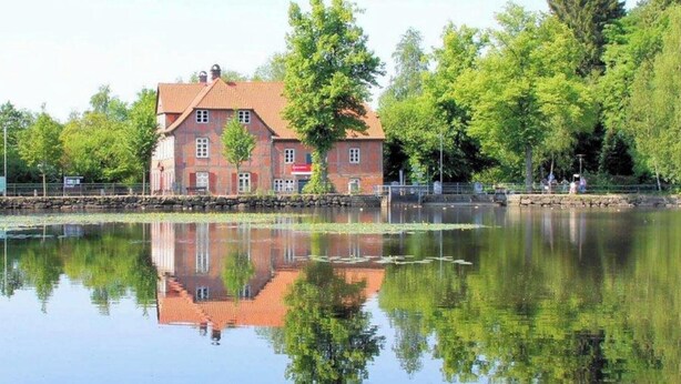 A red brick building with a tiled roof, reflecting in a calm body of water, surrounded by trees. - Urban & close to nature at Alter Bahnhof near Hamburg Bright apartment with 3 bedrooms (Trittau)