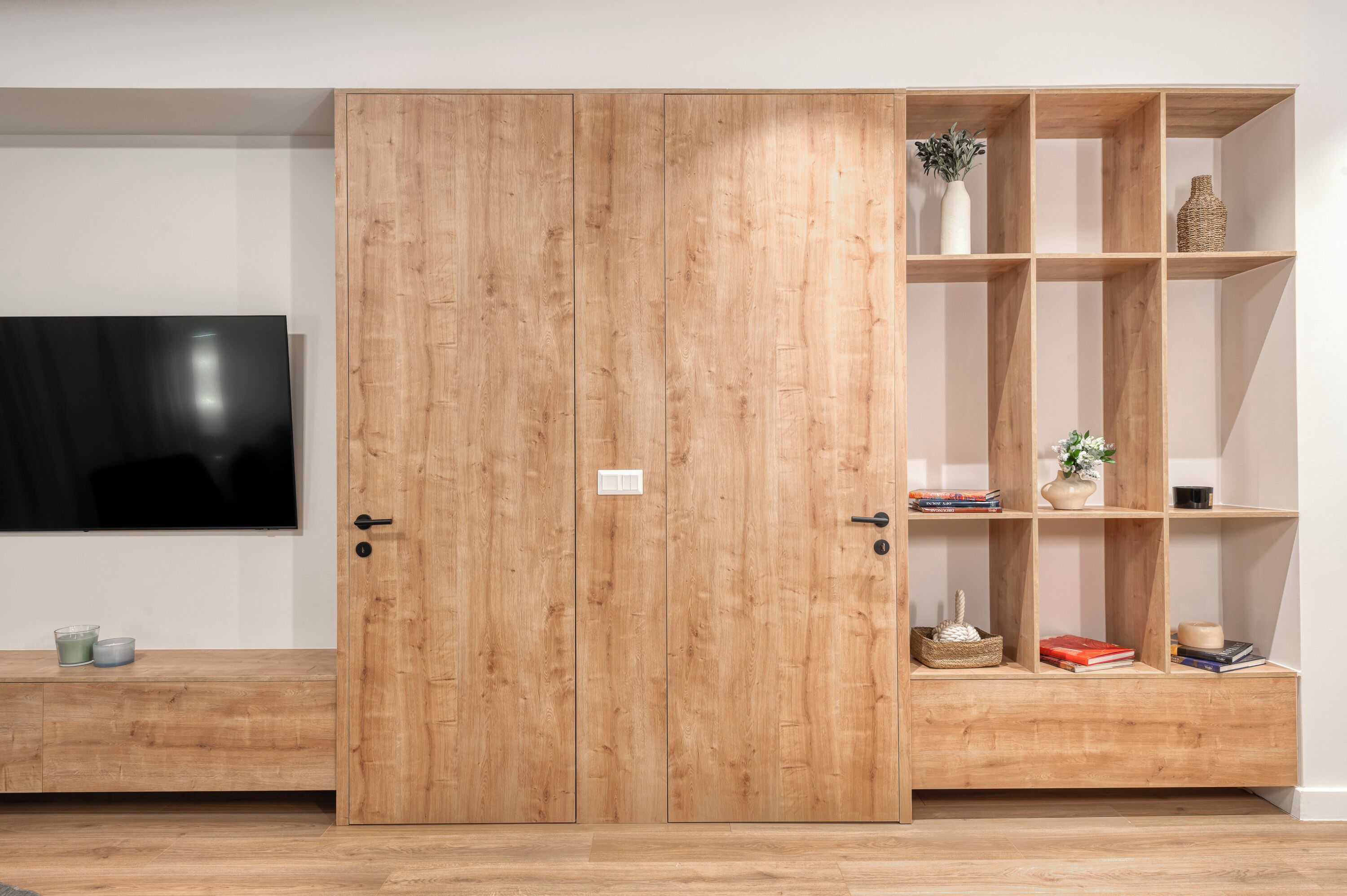 A wooden wardrobe with a TV on the wall, a shelf with books and decorative items, and a bench with candles.