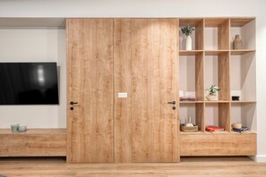 A wooden wardrobe with a TV on the wall, a shelf with books and decorative items, and a bench with candles. - Spacious Modern Apartment- Heart of Glyfada Center (Glifada)