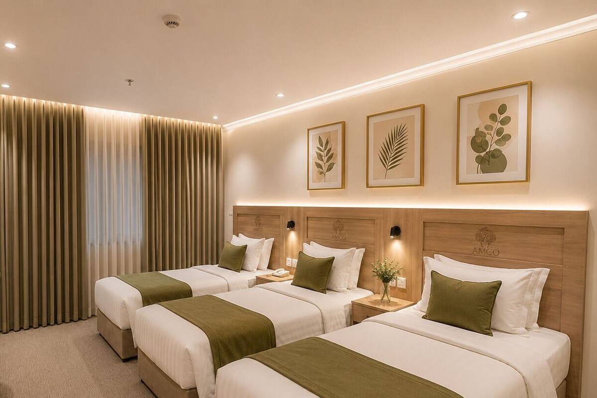 Habitación de hotel con tres camas, cabecera de madera, luces fijadas a la pared e impresiones botánicas enmarcadas.