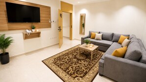 A modern living room with a grey sofa, a coffee table, a flat-screen TV, a wooden cabinet, a potted plant, and a patterned rug. - AMCO Serviced Apartments (Jeddah)