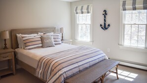 Un dormitorio con cama, mesitas de noche, un banco y dos ventanas con cortinas.