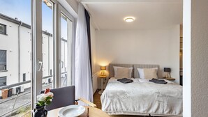 Een hotelkamer met een bed, nachtkastjes, een raam met gordijnen, een vaas met bloemen en uitzicht op gebouwen.