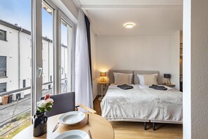 Camera d'albergo con un letto, comodini, una finestra con tende, un vaso con fiori e vista sugli edifici.